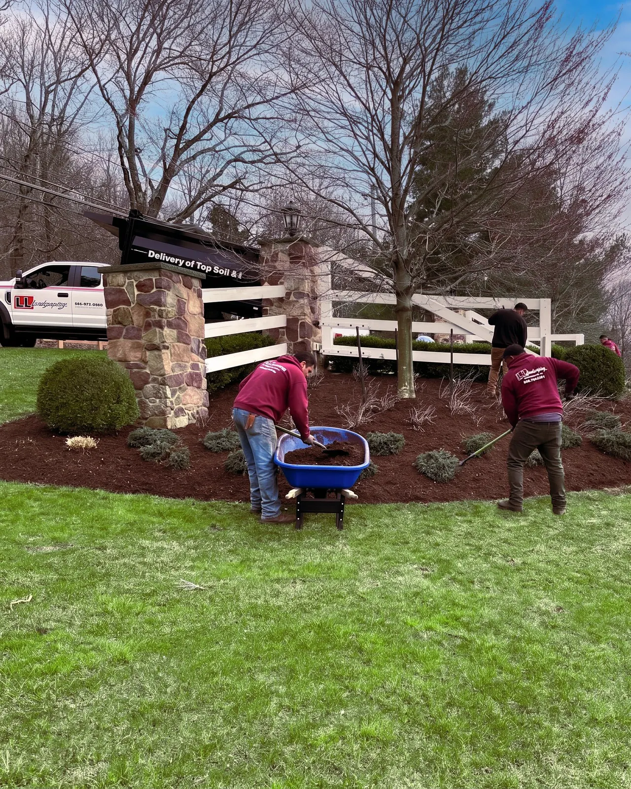 Two LL Landscaping crew members in red jackets working a curved mulch bed, one bending over a blue wheelbarrow, one raking, along a white split-rail fence, with the LL work truck and a stone-pillar gateway in the upper left.