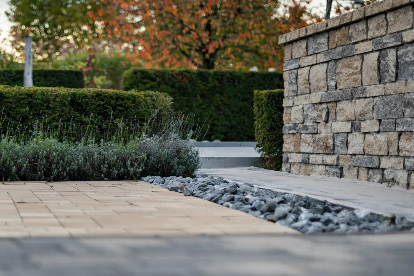 A finished paver patio with a river-rock drainage strip leading to a finely fitted stone column, trimmed boxwood hedge mid-frame, and warm autumn foliage in the background.
