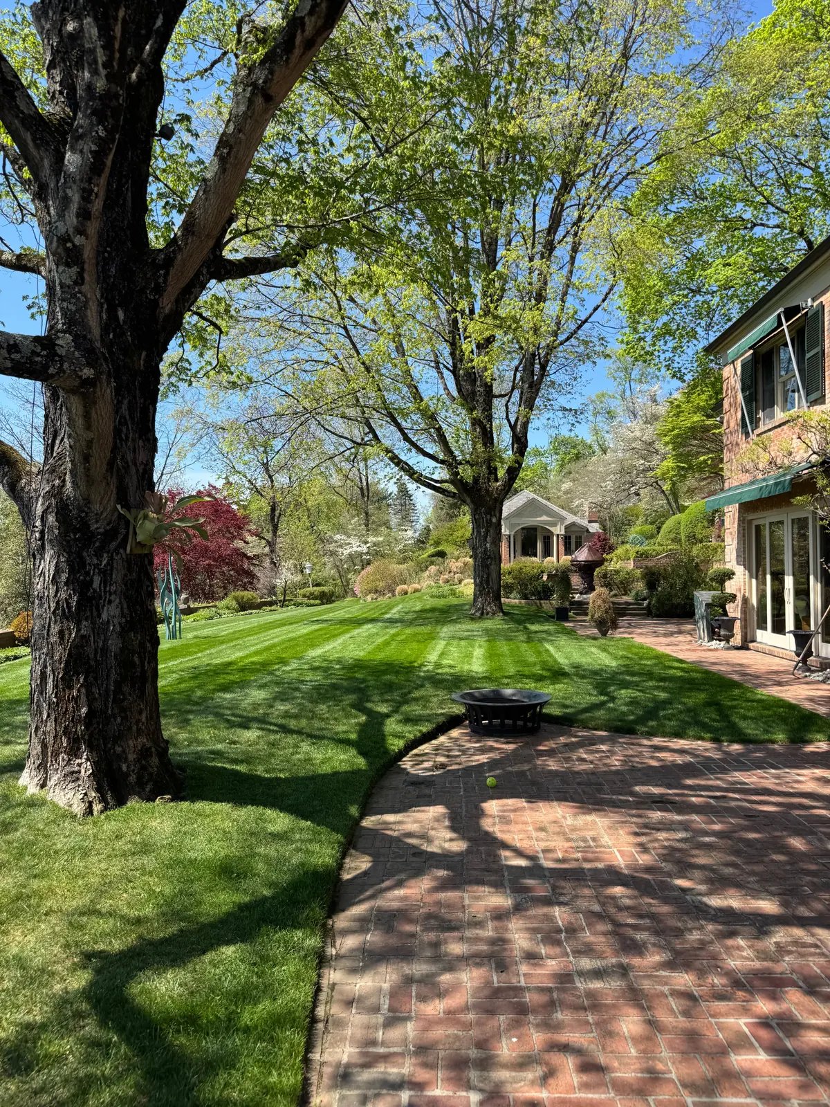 A manicured estate lawn with fresh mower stripes framed by mature shade trees, a curved brick walkway and patio, and flowering trees beside a brick home.