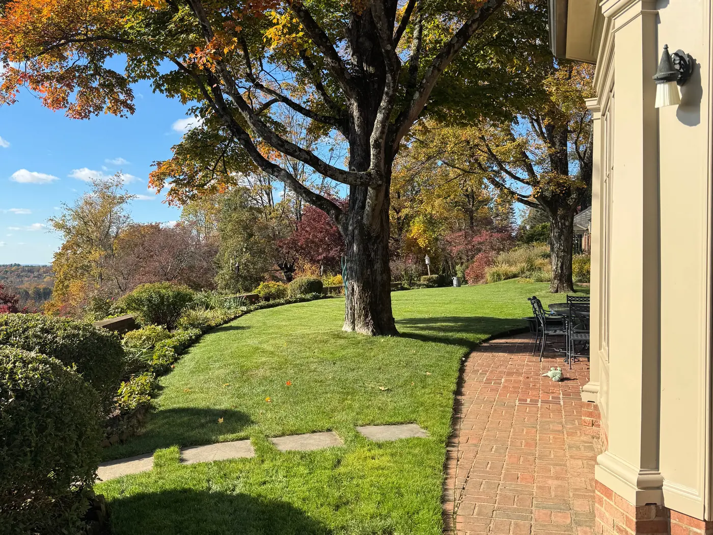 The same estate property after a fall cleanup, crisp green lawn cleared of leaves, a swept brick walkway, and the maple in full autumn color under a blue sky.