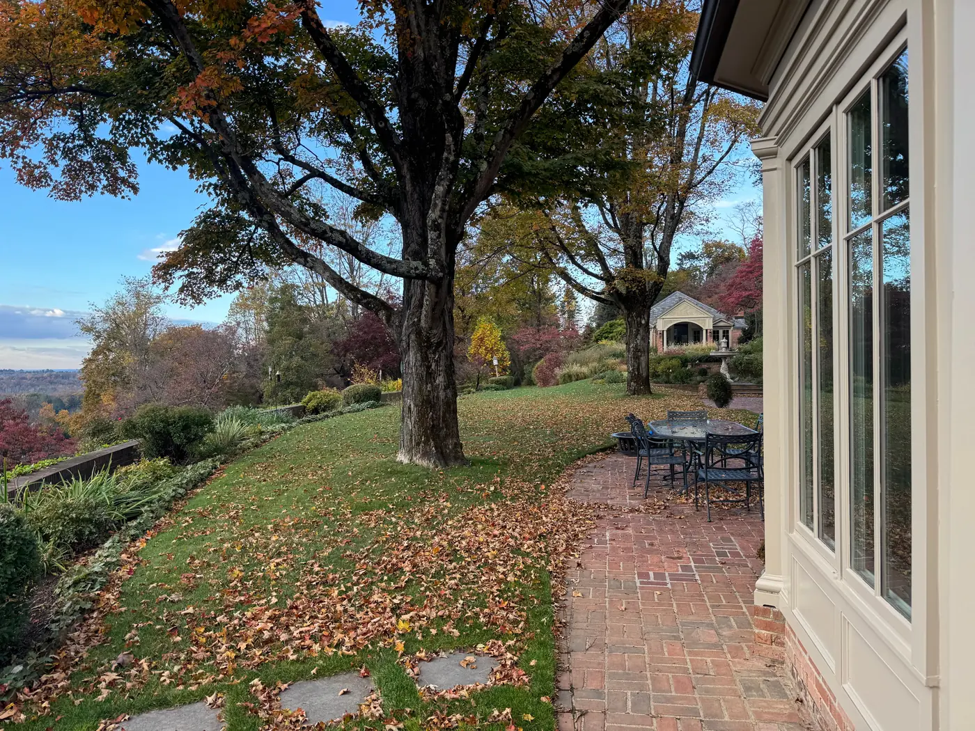 An estate lawn and brick patio blanketed with fallen autumn leaves before cleanup, a large maple in fall color overhead and a wooded valley beyond.