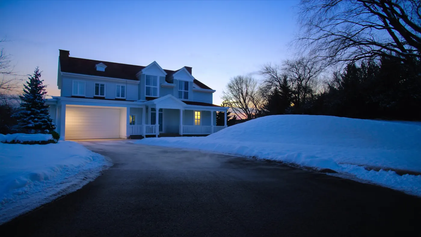 A residential driveway perfectly cleared of snow at blue-hour first light, leading up toward a white colonial home with the porch light glowing, plowed snow banks on both sides and mature trees flanking the property.
